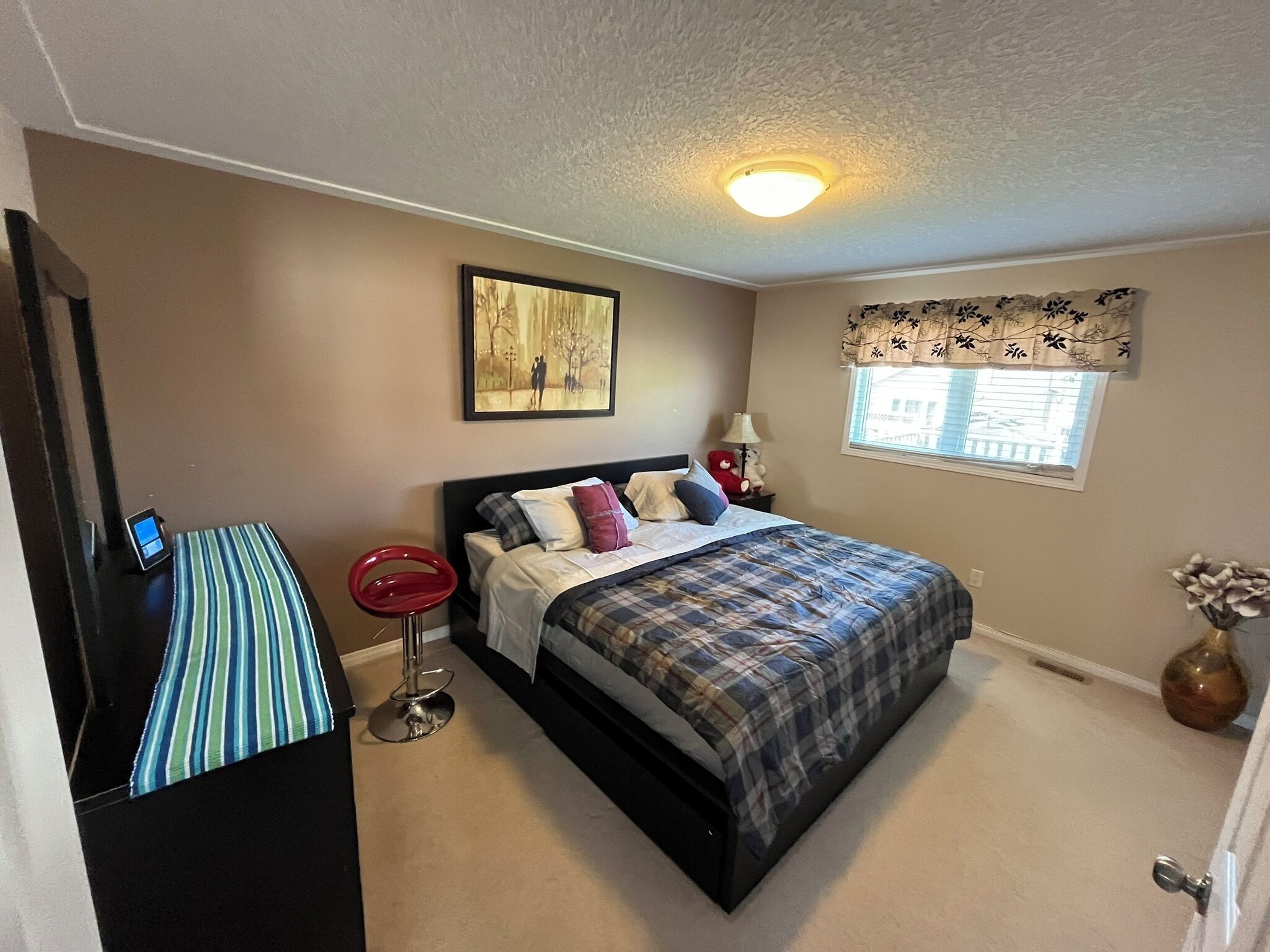 A bedroom with a bed, a bedside table with a lamp, a window with a view, and a painting on the wall.