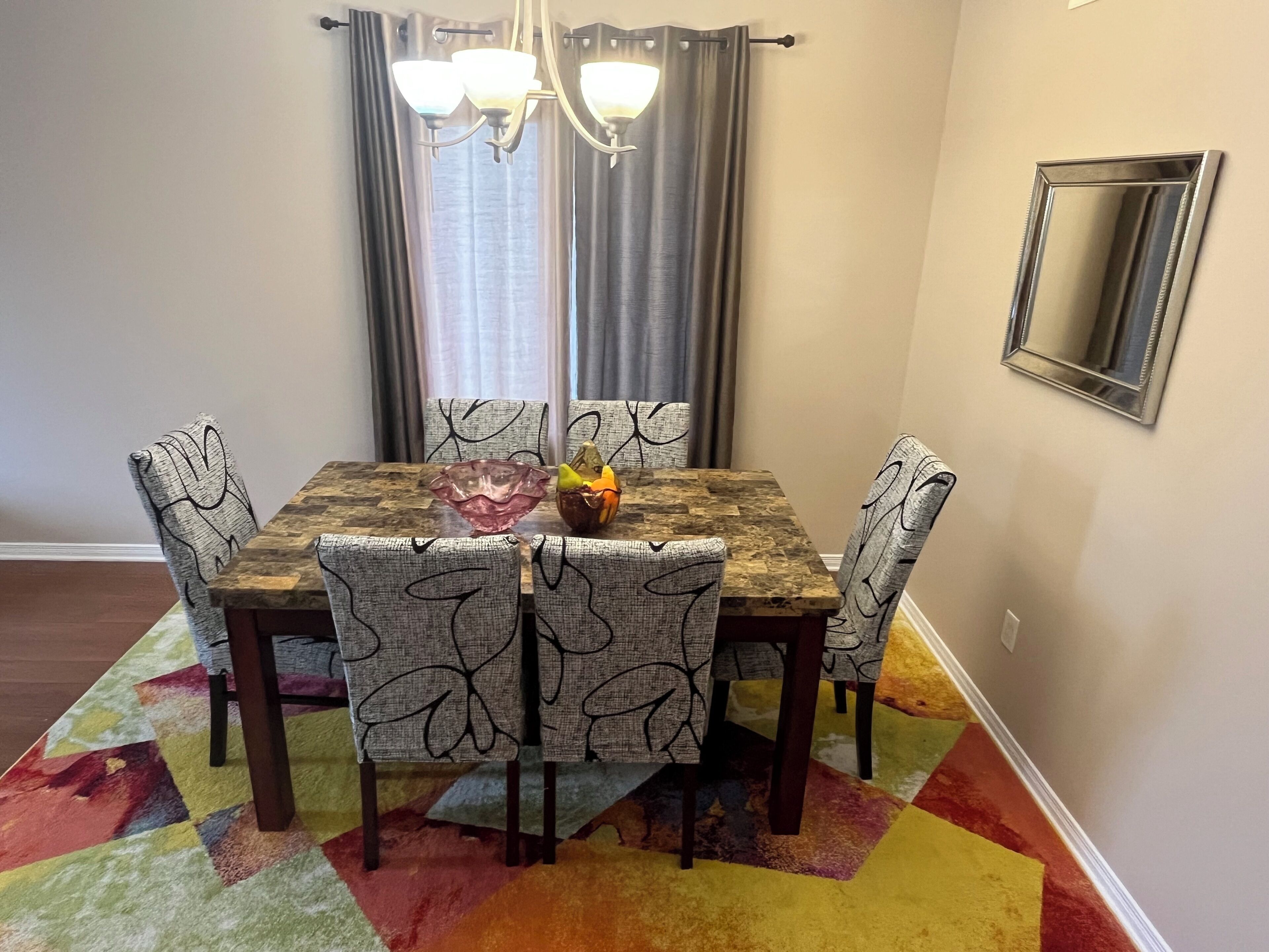 A dining area with a wooden table, four chairs with a leaf pattern, a fruit bowl, and a chandelier.
