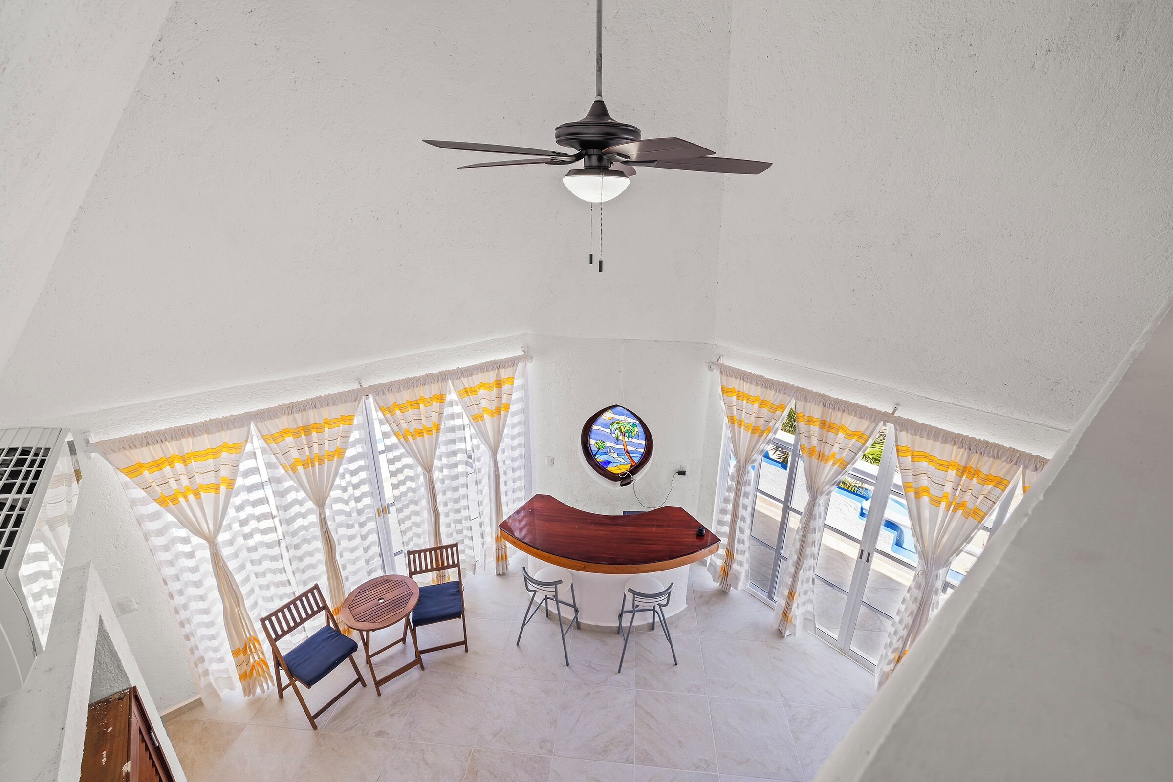 A dining area with a wooden table and chairs, a ceiling fan, and a view of the outdoors through large windows with curtains.