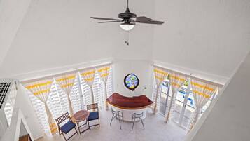 A dining area with a wooden table and chairs, a ceiling fan, and a view of the outdoors through large windows with curtains.