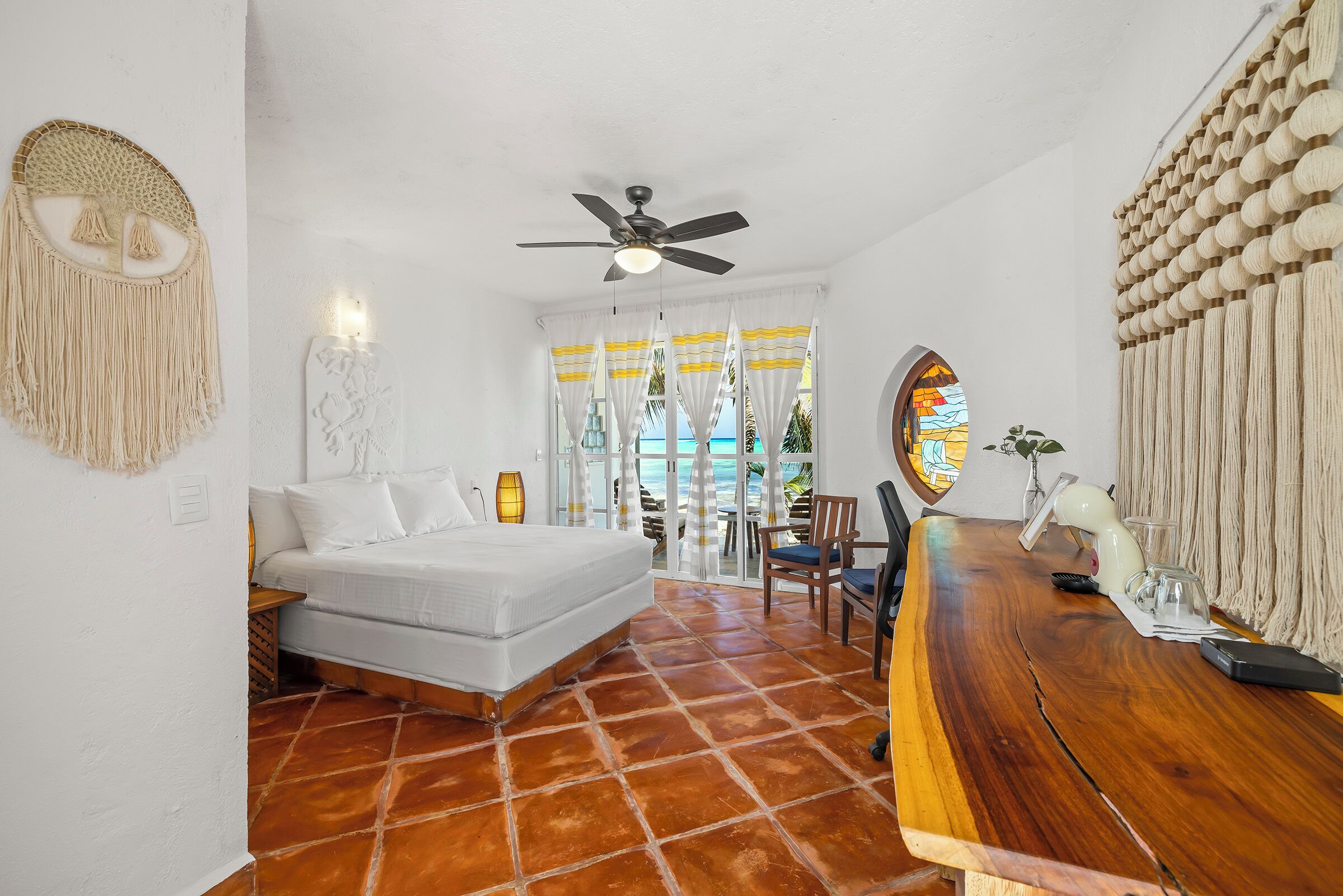 A bedroom with a bed, a ceiling fan, a wooden table, and a view of the sea through curtains.