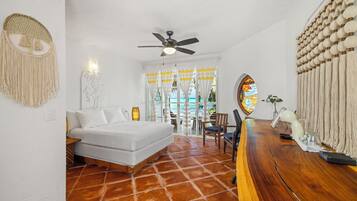 A bedroom with a bed, a ceiling fan, a wooden table, and a view of the sea through curtains.
