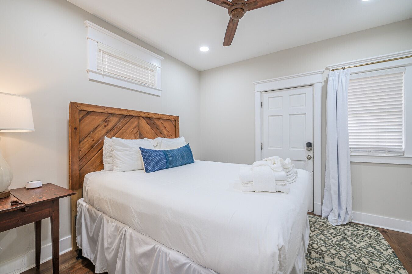 Un lit bien fait, avec une literie blanche et un oreiller bleu, une tête de lit en bois, une table de chevet avec une lampe et un ventilateur de plafond.