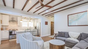 A spacious living room with a grey sofa, a striped armchair, and a round wooden coffee table. A kitchen with white cabinets and a built-in oven is visible in the background. - The Bogard Only 4 Blocks to King! Unit A (Charleston)