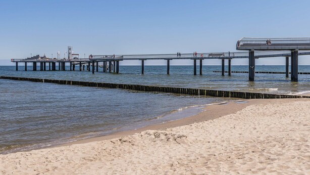 Une plage avec une jetée qui s’avance dans l’eau, dotée d’une promenade et où plusieurs personnes se promènent.