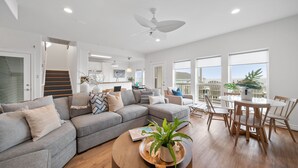 A modern living room with a grey sectional sofa, a wooden coffee table, and a dining area with wooden chairs and a white table. - Oceanfront 5BR Hot Tub & Swim Spa (Galveston)