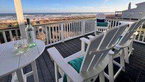 A deck with white chairs and a table, overlooking a beach with dunes and a building in the background. - Oceanfront 5BR Hot Tub & Swim Spa (Galveston)