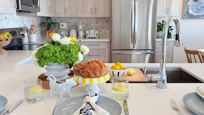 A modern kitchen with a stainless steel refrigerator, a microwave, and a sink. There is a vase with flowers on the counter, a plate with a napkin, and a glass of water. - Oceanfront 5BR Hot Tub & Swim Spa (Galveston)