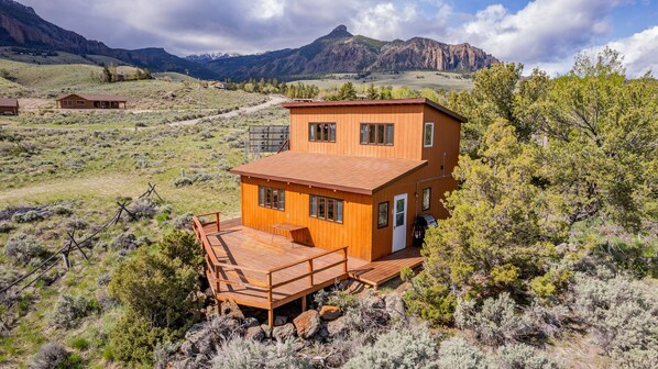 A wooden cabin with a deck, surrounded by mountains and sparse vegetation. - Loren's Lookout- pet friendly, on the way to Yellowstone National Park (Cody)