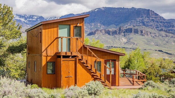 A wooden cabin with a balcony, a staircase, and a deck, surrounded by mountains and vegetation. - Loren's Lookout- pet friendly, on the way to Yellowstone National Park (Cody)