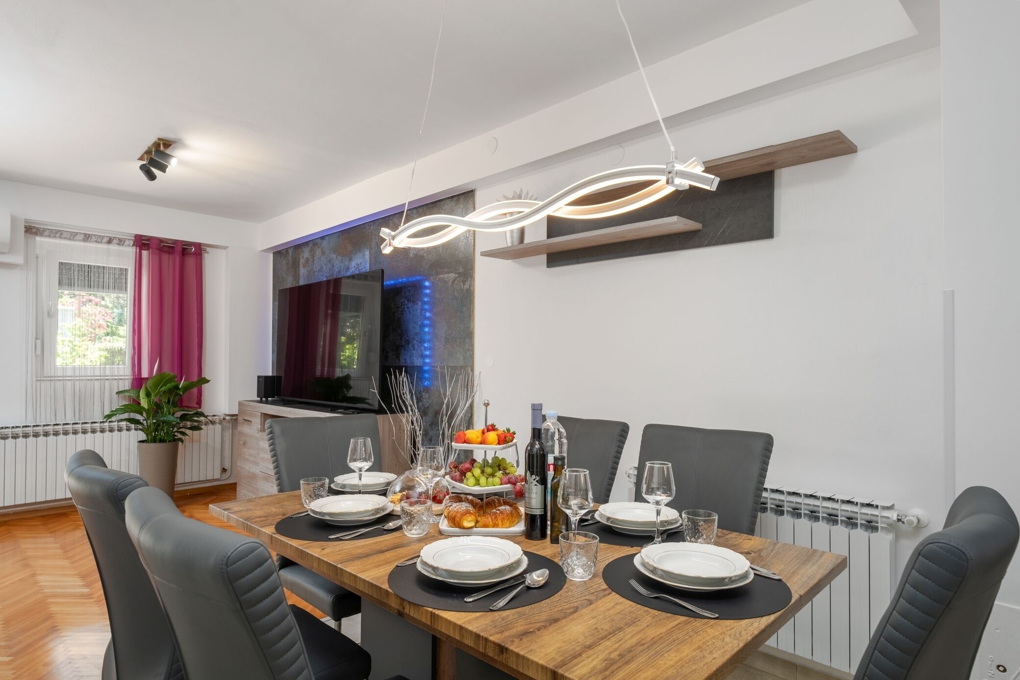 A dining area with a wooden table set for four, surrounded by black chairs. A fruit platter and a bottle of wine are on the table.
