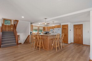A spacious kitchen with a central island, wooden bar stools, and a staircase leading to an upper level. - Cloudbreak: Pet Friendly, Oceanside, Hot Tub, Community Amenities! (Duck)