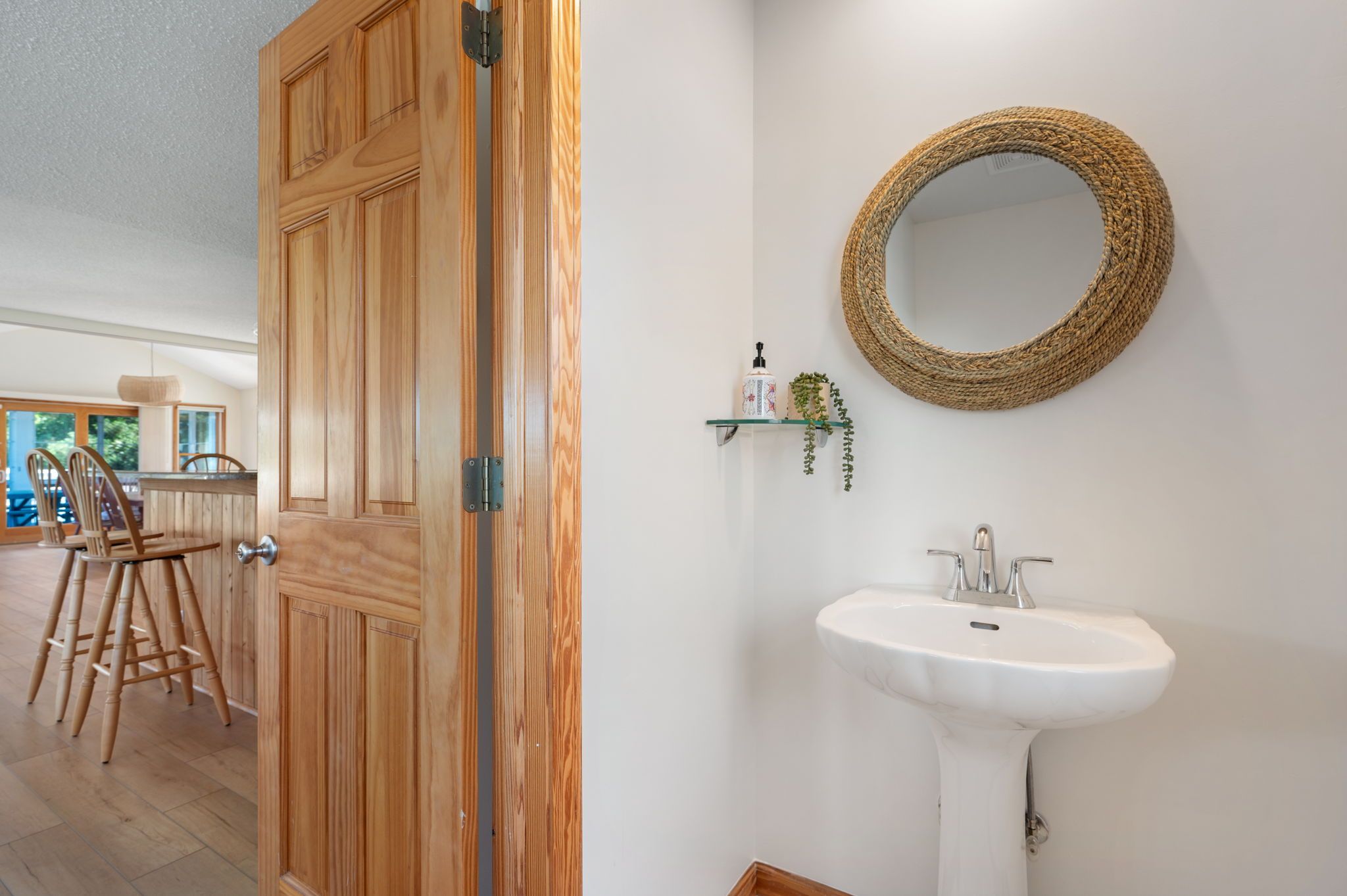 Une porte en bois dotée d’une poignée moderne, un grand miroir rond fixé au mur et peint en blanc, un lavabo sur pied blanc et une petite étagère avec une plante et une bouteille.