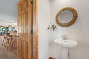 A wooden door with a modern handle, a white wall-mounted round mirror, a white pedestal sink, and a small shelf with a plant and a bottle. - Cloudbreak: Pet Friendly, Oceanside, Hot Tub, Community Amenities! (Duck)