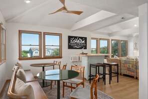A spacious living area with a dining table, chairs, a sofa, and a colorful patterned couch. There are large windows providing views of the outside.