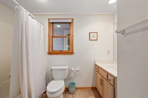 Een badkamer met een toilet, douchegordijn, raam, wastafel en kastje.