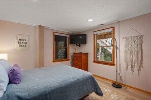 A bedroom with a bed, a dresser, a TV, a macrame wall hanging, and a window with a view of a wooden deck and stairs. - Cloudbreak: Pet Friendly, Oceanside, Hot Tub, Community Amenities! (Duck)