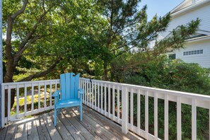 A wooden deck with a blue Adirondack chair, surrounded by white railings and lush greenery.