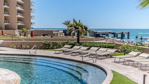 A pool area with lounge chairs, a palm tree, and a view of the ocean. - Las Palomas Opalo 1003 2BR 2BA Unit. (Puerto Peñasco)