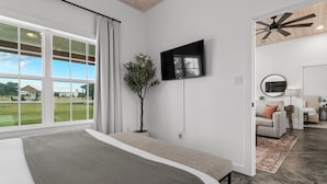 Un dormitorio moderno con un ventanal grande, un televisor de pantalla plana, un ventilador de techo y vistas a un paisaje verde.