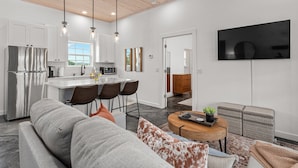 Una cocina moderna con electrodomésticos de acero inoxidable, un comedor con mesa redonda y una sala de estar con sofá y televisor montado en la pared.
