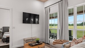 A modern living room with a flat-screen TV, a round coffee table, a sofa, and a large window offering a view of a green landscape. - Cross Mountain Cottages 7 | Ensuite Baths & Porch (Fredericksburg)