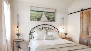 Une chambre à coucher avec un lit en métal, du linge de lit blanc, un tableau représentant un chemin de campagne et deux lampes de chevet.