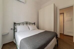 A single bed with a metal frame, white bedding, and a gray blanket. There is a white wall with a power outlet and a door to the right. - Testaccio 11 - FromHometoRome (Roma)