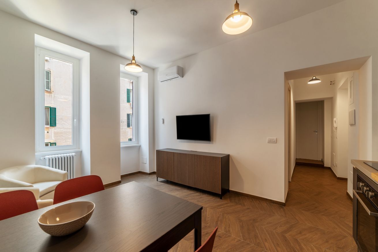 A modern living room with a dining area, a TV mounted on the wall, and a kitchen visible through an open doorway.