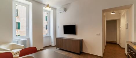 A modern living room with a dining area, a TV mounted on the wall, and a kitchen visible through an open doorway.