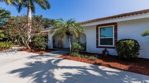 A single-story house with a tiled roof, a front porch, and a small garden with palm trees and flowering plants. - As you approach this lovely vacation home, lush greenery and shade trees greet you. (Marco Island)
