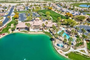 An aerial view of a resort with a large pool, multiple buildings, and a surrounding landscape with greenery and a body of water.
