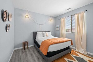 A bedroom with a bed, a small stool, a wall-mounted light fixture, and a window with curtains.