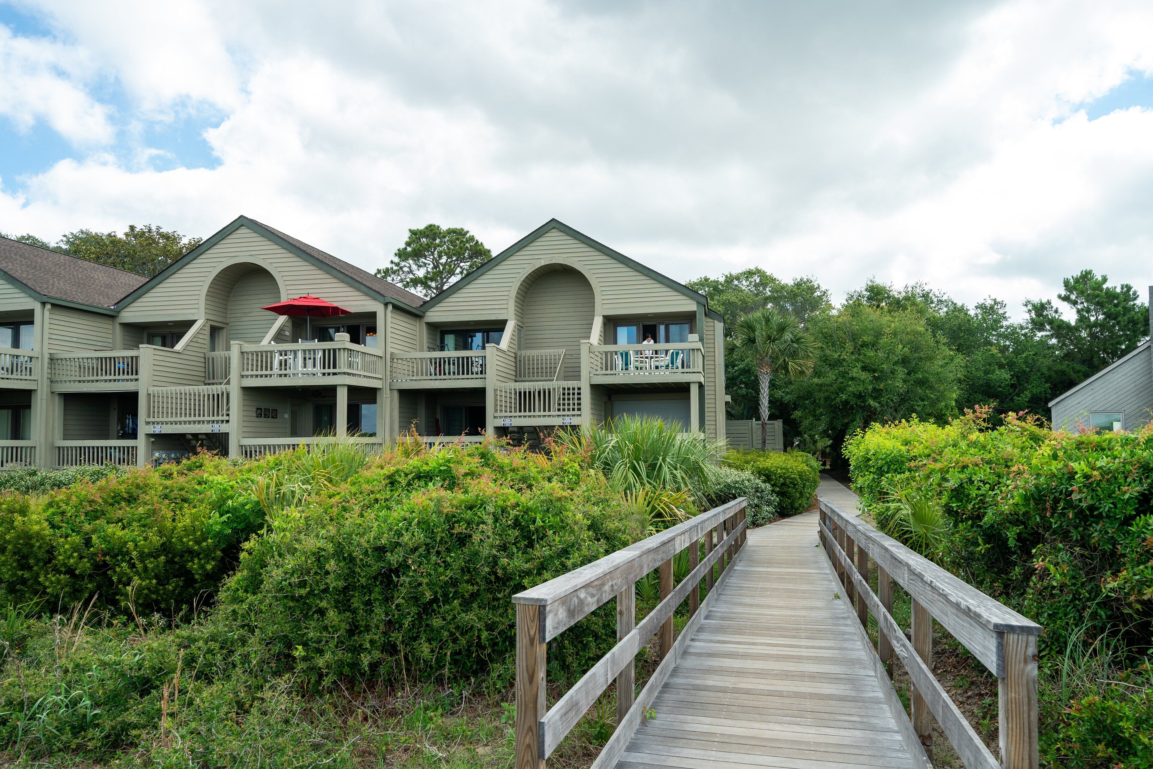 A wooden boardwalk leading to a row of two-story buildings with balconies and a red umbrella.