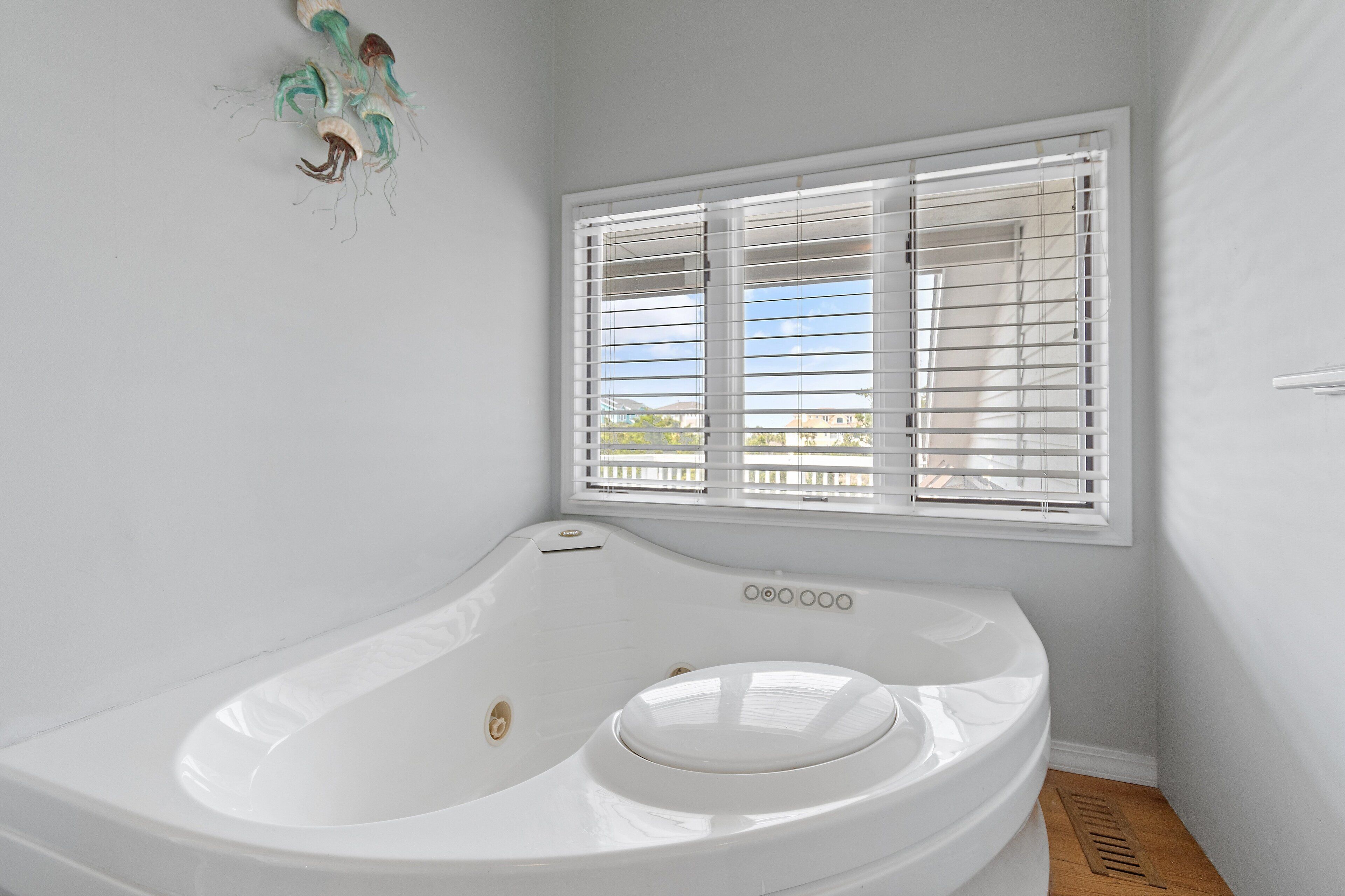 A bathroom with a white bathtub, a window with blinds, and a wooden floor.