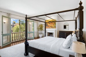 A four-poster bed with a white bedspread, a nightstand with a lamp, a fireplace, and a balcony with a view of trees and buildings. - The Canal Escape - canal-front Italian villa (Los Angeles)
