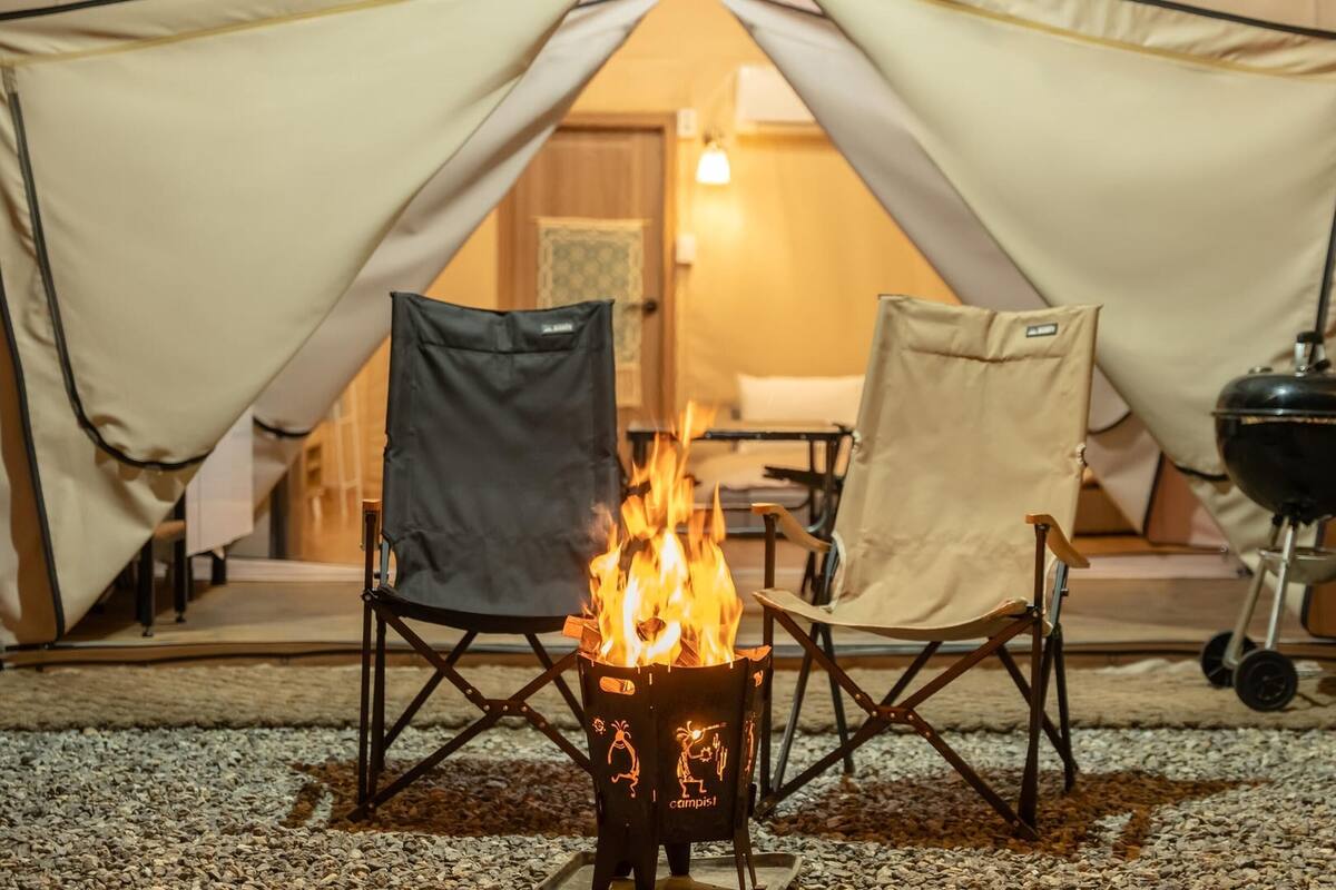 A tent with a campfire in the center, surrounded by camping chairs and a grill.