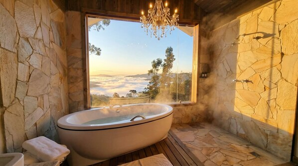 Une salle de bain aux murs en pierre, dotée d’une baignoire sur pieds, d’une grande fenêtre et d’un lustre.