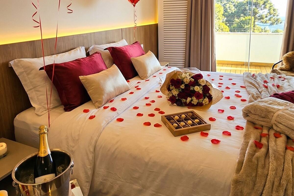 A hotel room with a bed, a bedside table with a bottle of champagne, a bouquet of flowers, and heart-shaped balloons.