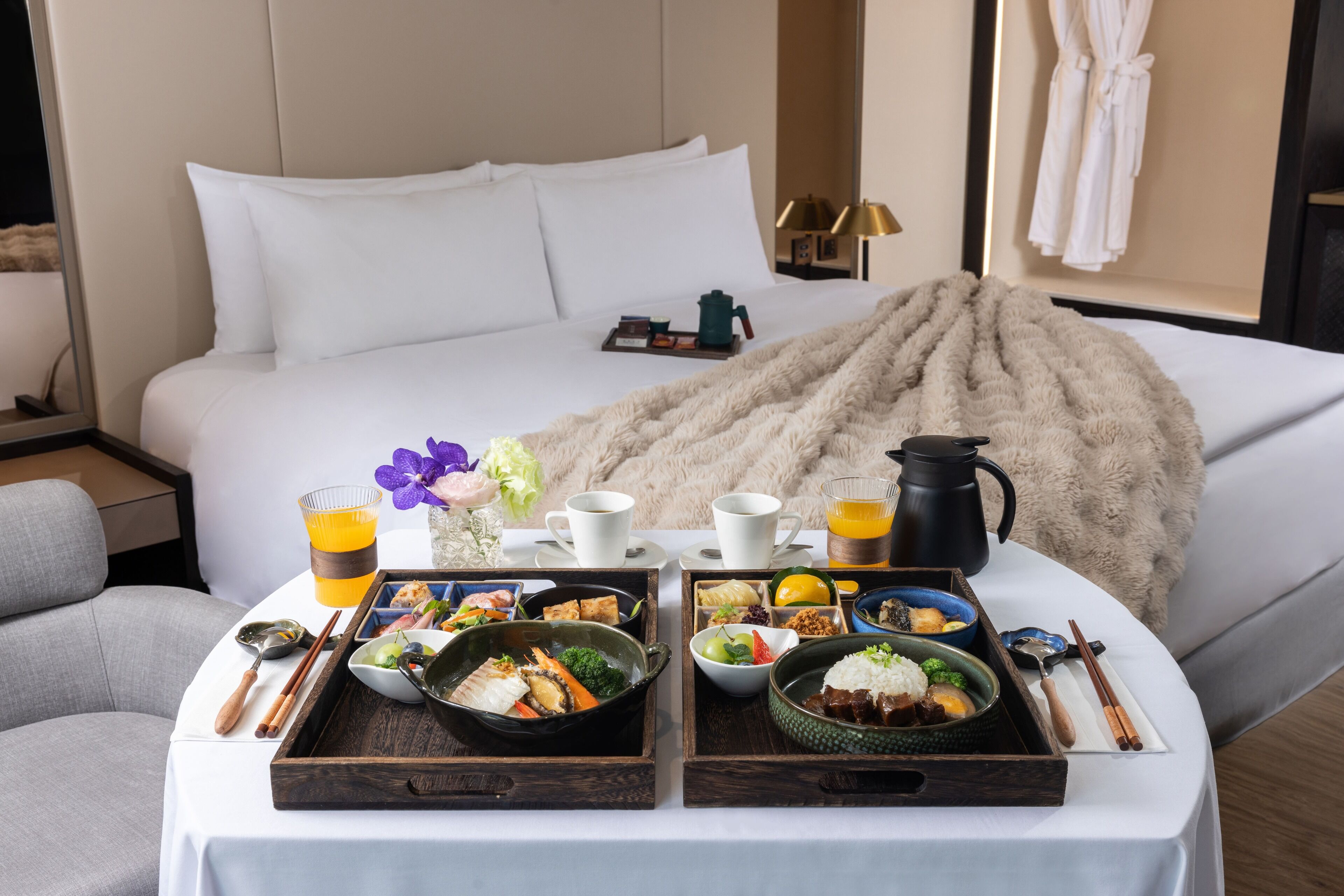 A neatly arranged breakfast buffet on a hotel bed with a fluffy blanket, a coffee pot, and a tray of food including rice, vegetables, and soup.