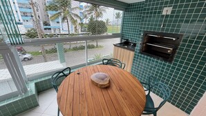 Een balkon met een houten tafel en stoelen, een kitchenette met een magnetron, en uitzicht op een straat met gebouwen en palmbomen.