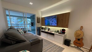 Een moderne woonkamer met een grijze bank, een salontafel met een boek en afstandsbediening, een keukenhoek met een televisie, en een gitaar aan de muur.