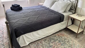 Una cama bien hecha con un edredón oscuro y almohadas blancas, una mesita de noche con una lámpara y una alfombra estampada sobre piso de madera.