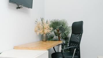 Ein kleiner Büroraum mit einem weißen Schreibtisch, einem schwarzen Bürostuhl, einem wandmontierten Flachbildfernseher und einem Blumentopf.