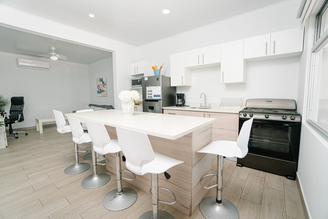 Een moderne keuken met een centraal eiland, witte barkrukken, een zwarte oven en witte kasten.