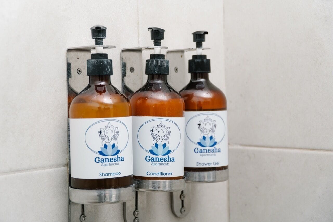 Trois flacons de produits Ganesha : un shampoing, un après-shampoing et un gel douche, fixés au mur.