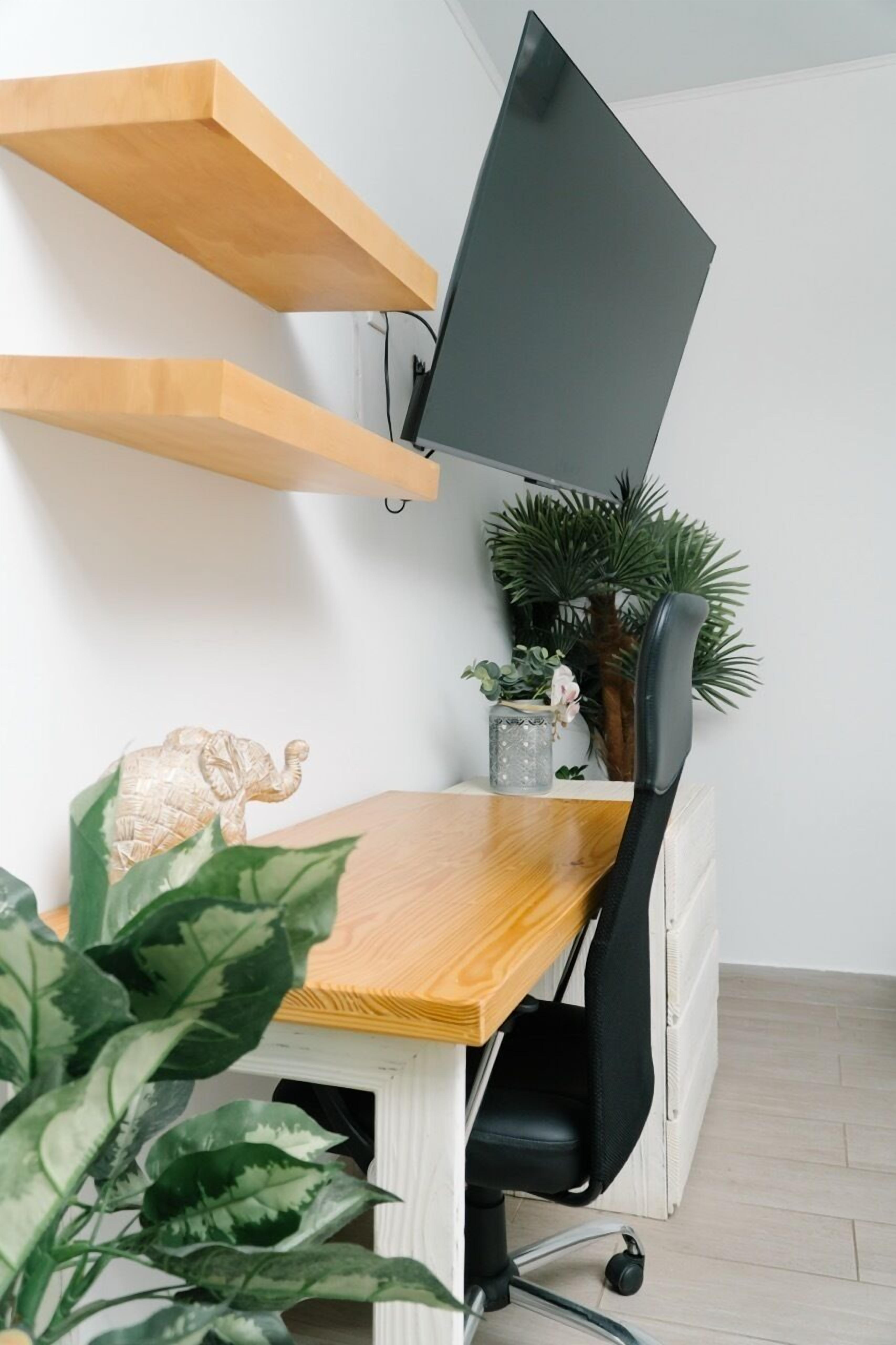Ett modernt kontorsutrymme med ett skrivbord i trä, en svart kontorsstol och en väggmonterad platt-TV. På skrivbordet står krukväxter och en dekorativ elefantfigur.