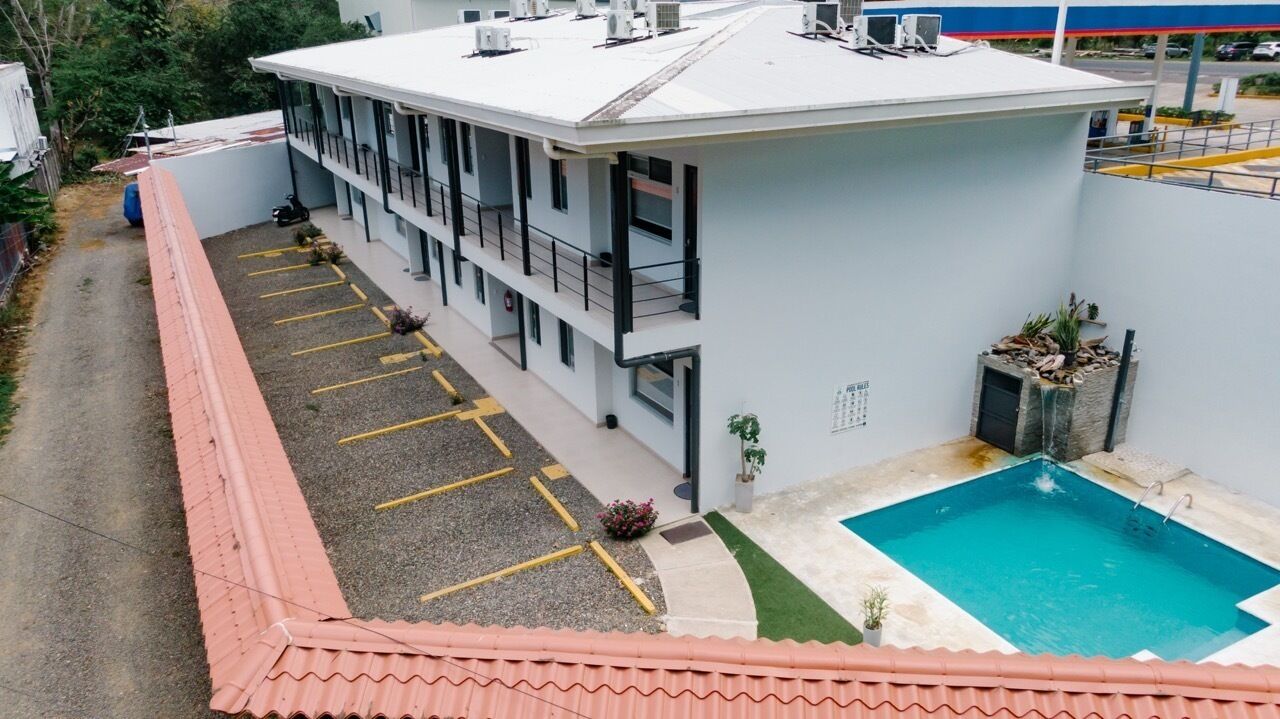 A building with a swimming pool, a parking area, and a red-roofed adjacent structure.