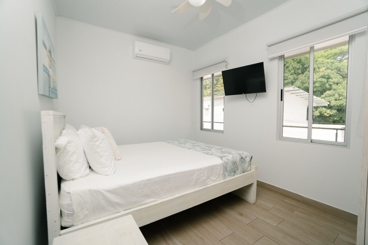 A bedroom with a bed, a TV mounted on the wall, and a window with a view of greenery.