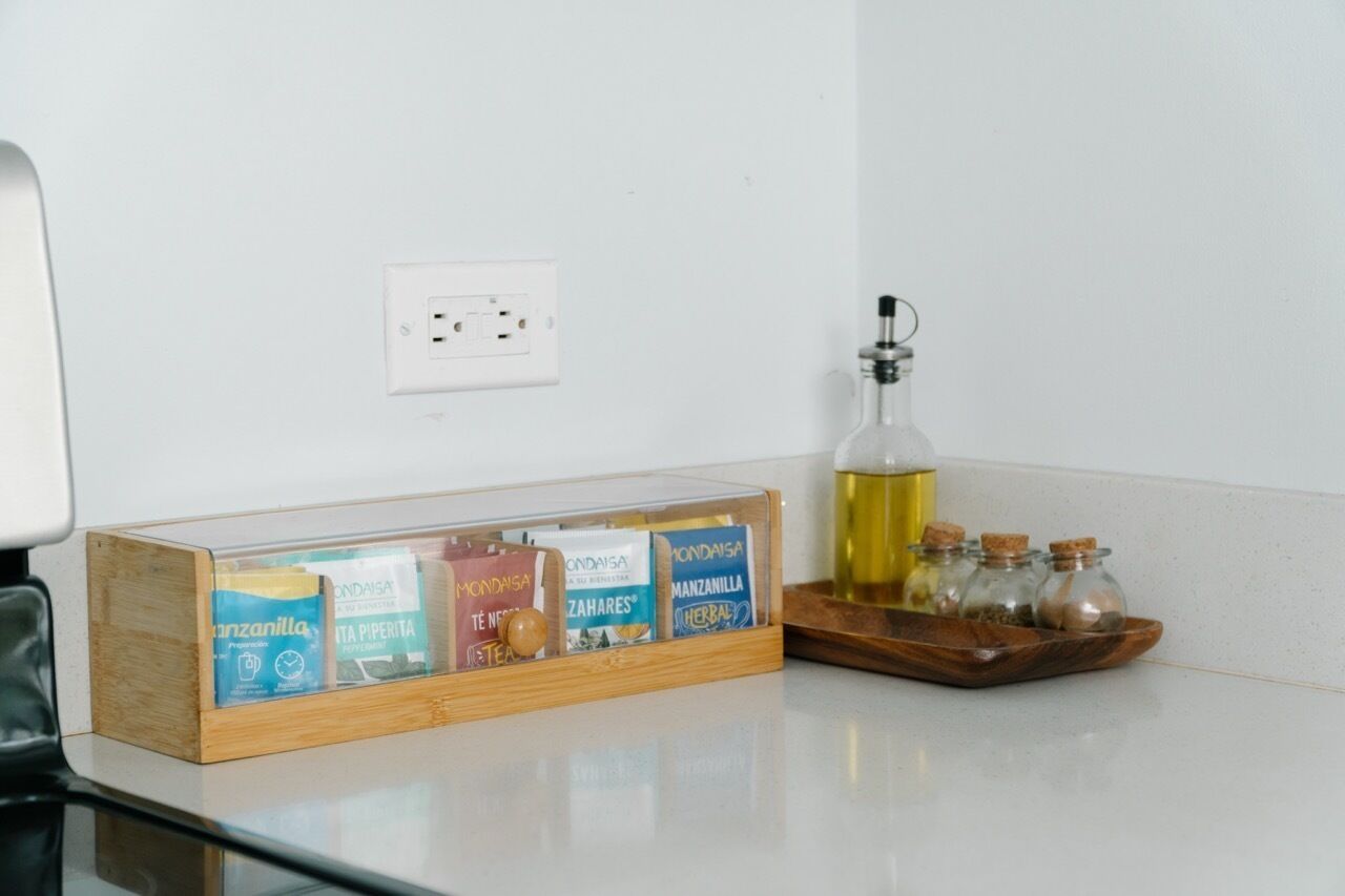 A kitchen counter with a wooden box containing tea bags, a bottle of oil, and small glass jars with spices.